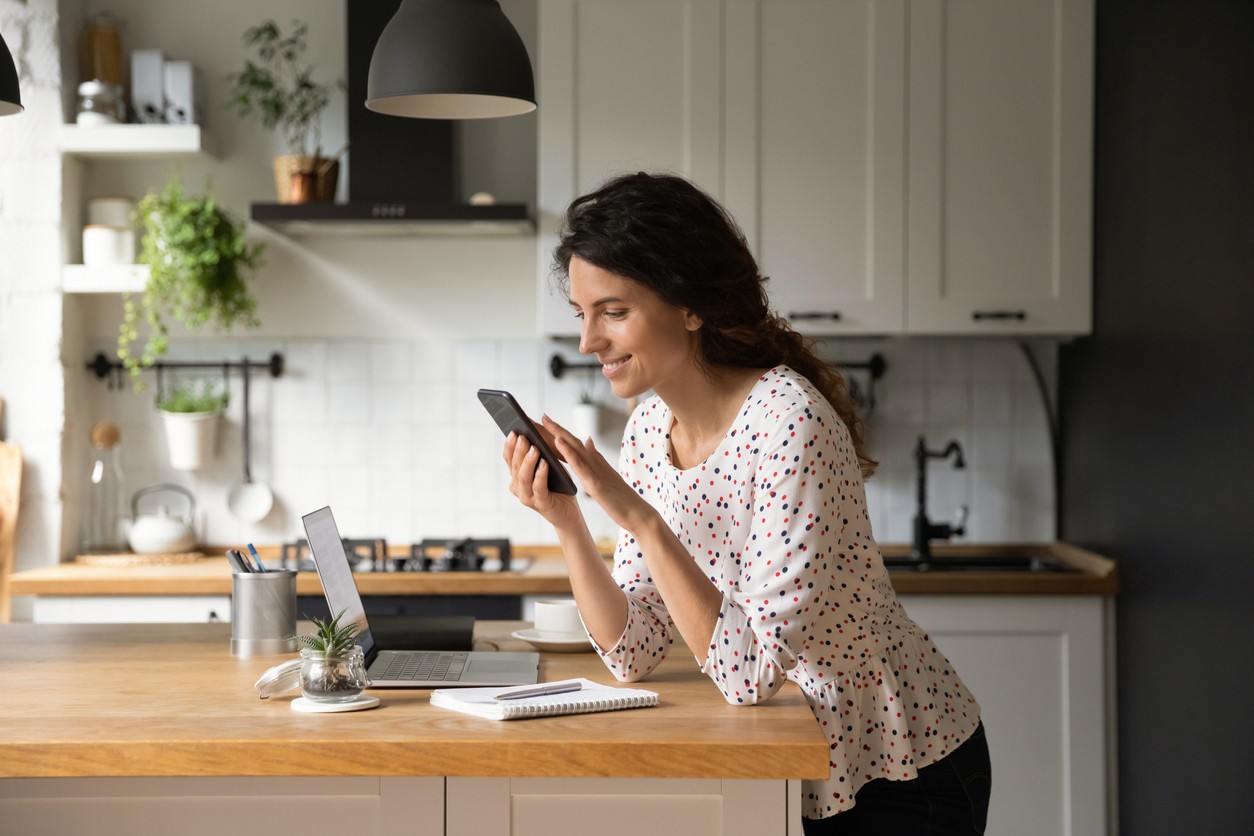 Happy woman looking at her phone in the kitchen with laptop, notebook and pen on top of the table